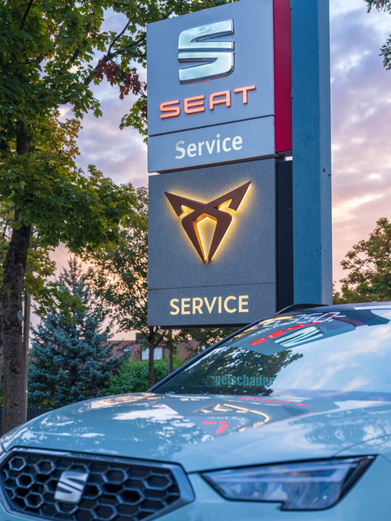 Beleuchtetes Schild mit den Logos für SEAT Service und CUPRA Service beim Autohaus Spratter in München, im Vordergrund die Frontpartie eines grauen SEAT-Fahrzeugs.