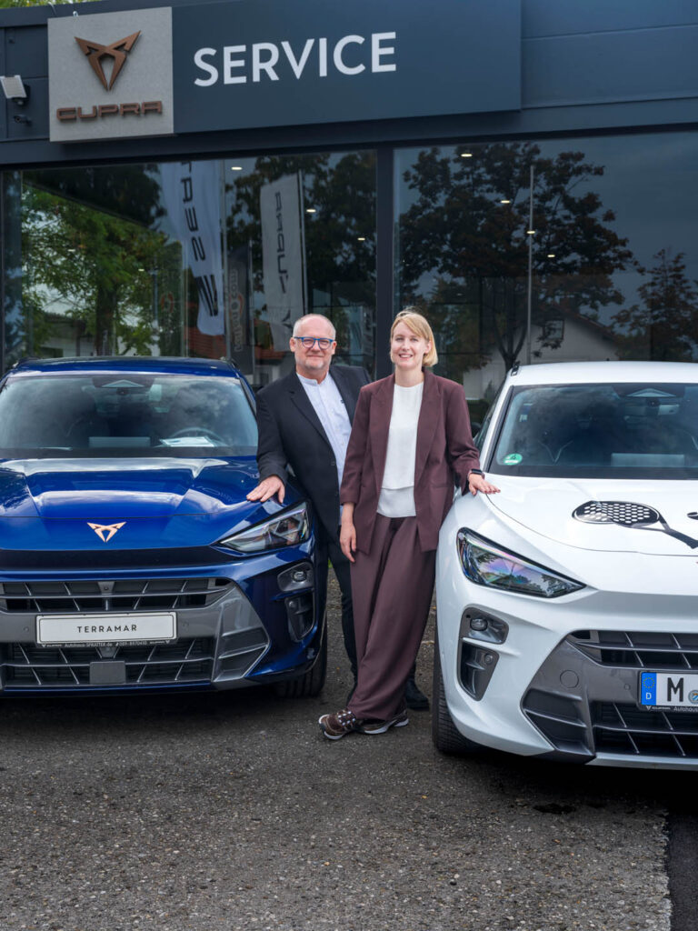 Helmut Spratter und Laura Hammers stehen zwischen zwei Autos von Cupra, vor dem Autohaus Spratter, unter dem Schild Cupra Service