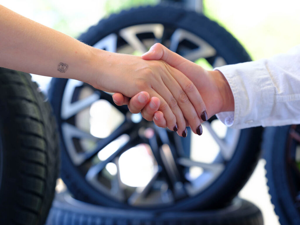 Nahaufnahme eines Handschlags zwischen Kunde und Mitarbeiter vor gestapelten Autoreifen mit Alufelgen, stellvertretend für zuverlässigen Service beim SEAT und CUPRA Partner Autohaus Spratter in München.
