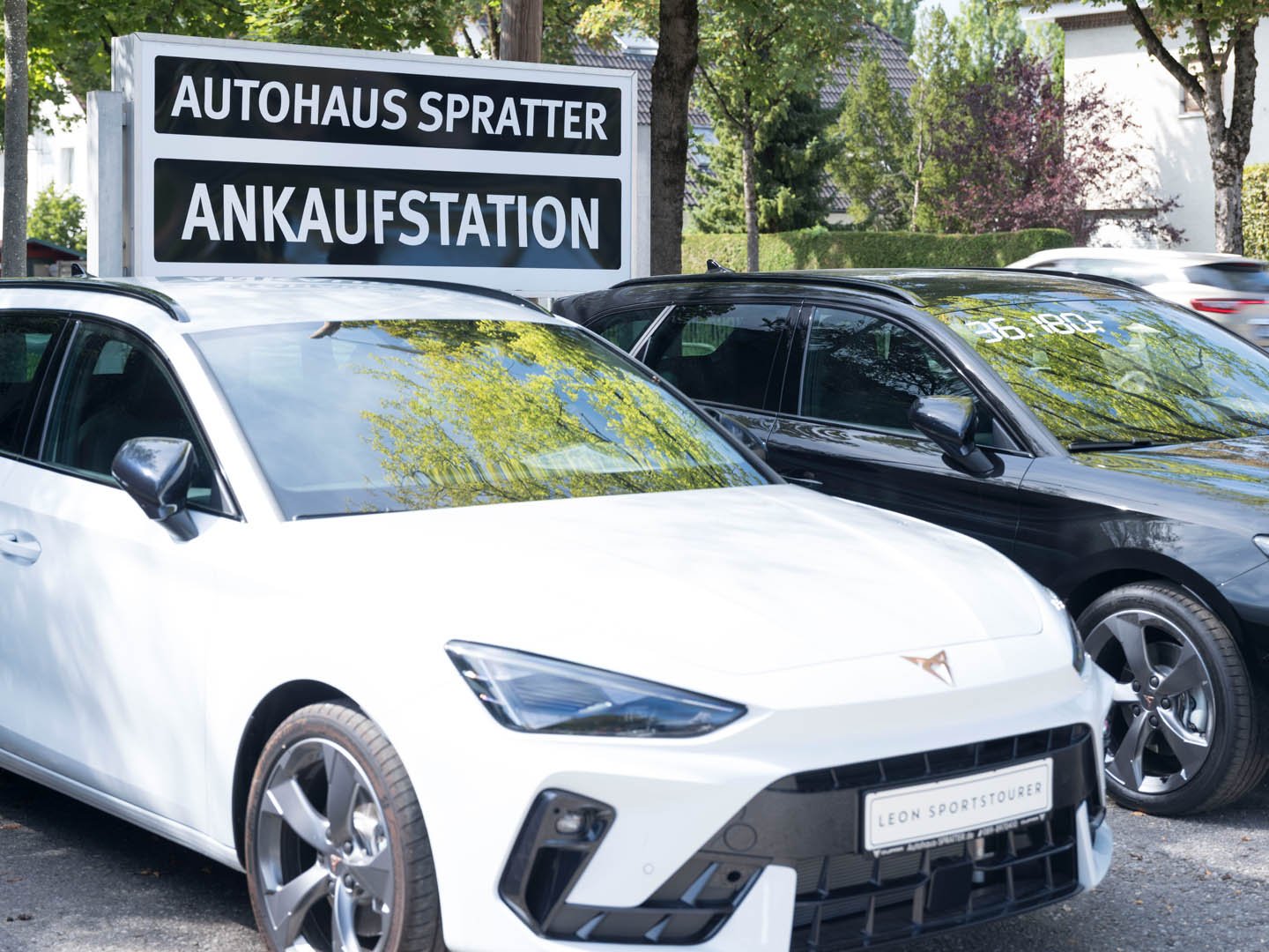 Unter dem Schild mit der Aufschrift AUTOHAUS SPRATTER ANKAUFSTATION steht ein weisses Auto vom Modell CUPRA Terramar und daneben steht ein graues Auto von SEAT