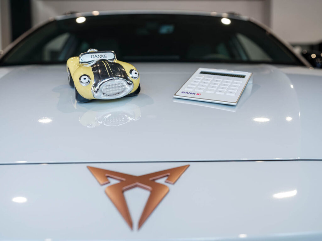 Eine Autoförmige Spardose und ein Taschenrechner liegen auf der Motorhaube eines weissen Autos