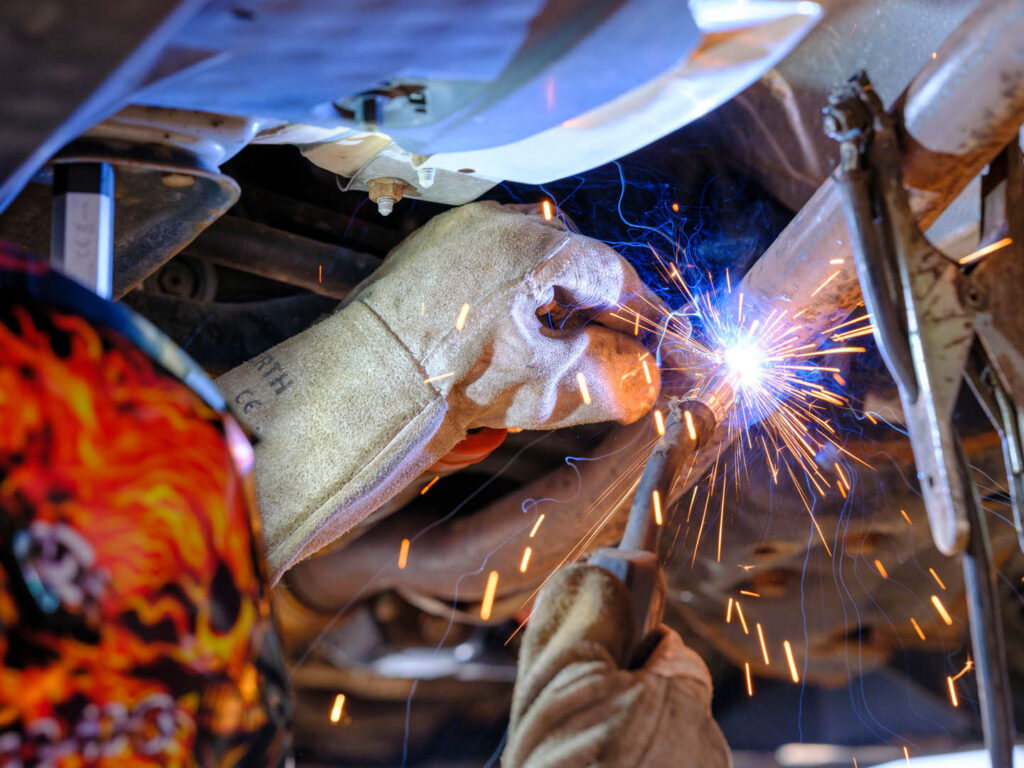 Ein KFZ-Mechaniker mit Schutzhandschuhen führt präzise Schweißarbeiten an der Unterseite eines Fahrzeugs in der Werkstatt von Autohaus Spratter (SEAT & CUPRA Service Partner in München) durch, mit hellem Funkenflug.