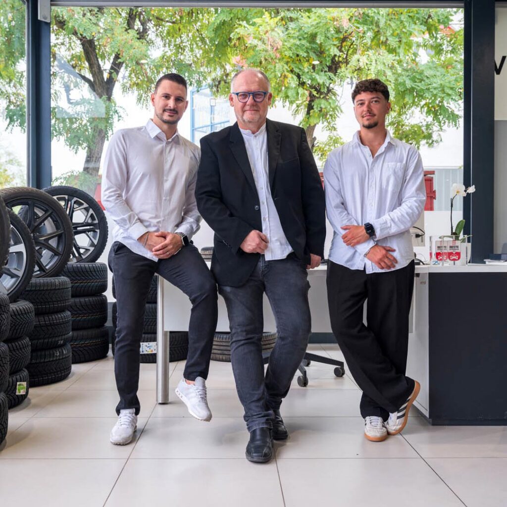 3 Mitarbeiter des Autohaus Spratter aus der Verkaufs-Abteilung stehen im Ausstellungsraum vor einem Schreibtisch daneben stehen mehrere Autoreifen im Hintergrund ist ein Baum zu sehen